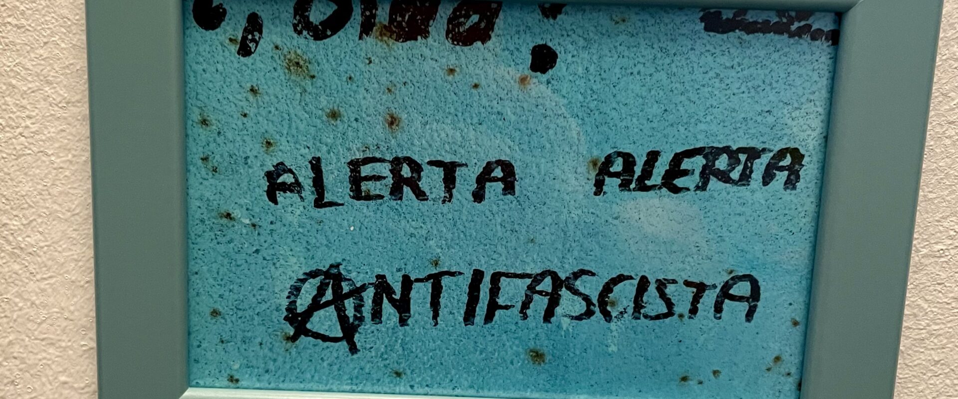 Ein gerahmtes Bild an einer weißen Wand zeigt den Schriftzug „ALERTA ALERTA ANTIFASCISTA“ in schwarzer Schrift auf einem türkisfarbenen Hintergrund. Das Wort „ANTIFASCISTA“ ist mit einem stilisierten „A“ im Kreis geschrieben, ein Symbol, das häufig mit Anarchismus assoziiert wird. Der Rahmen des Bildes ist helltürkisfarben. Im Hintergrund sind leichte Flecken oder Strukturierungen erkennbar.