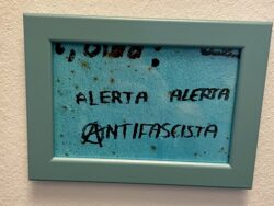 Ein gerahmtes Bild an einer weißen Wand zeigt den Schriftzug „ALERTA ALERTA ANTIFASCISTA“ in schwarzer Schrift auf einem türkisfarbenen Hintergrund. Das Wort „ANTIFASCISTA“ ist mit einem stilisierten „A“ im Kreis geschrieben, ein Symbol, das häufig mit Anarchismus assoziiert wird. Der Rahmen des Bildes ist helltürkisfarben. Im Hintergrund sind leichte Flecken oder Strukturierungen erkennbar.