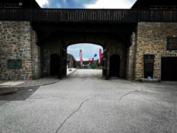 Ein steinernes Eingangstor des ehemaligen Konzentrationslagers Mauthausen mit einem dunklen Holzüberbau. Der Torbogen ist aus grob behauenem Naturstein gemauert und führt durch einen Tunnelgang. Durch den Torbogen hindurch sind rote Fahnen oder Banner zu erkennen. Der Himmel zeigt dramatische Wolkenformationen. Der Boden besteht aus rissigem Asphalt. Die Aufnahme vermittelt durch die Perspektive und den Kontrast zwischen dem dunklen Tor und dem hellen Himmel eine bedrückende Atmosphäre.