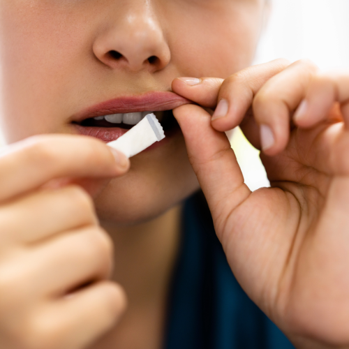 Nahaufnahme einer Person, die einen Nikotinbeutel mit den Fingern an ihre Oberlippe führt. Die Lippen sind leicht geöffnet, und die Hände halten den kleinen, weißen Beutel vorsichtig fest. Im Hintergrund ist ein unscharfer, heller Bereich zu sehen.