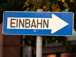 Blaues Verkehrsschild mit weißem Pfeil nach rechts und dem schwarzen Schriftzug „Einbahn“ in Großbuchstaben. Im Hintergrund sind unscharf Bäume mit herbstlichen Blättern sowie ein Gebäude zu sehen.