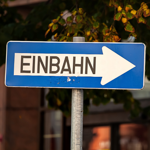 Blaues Verkehrsschild mit weißem Pfeil nach rechts und dem schwarzen Schriftzug „Einbahn“ in Großbuchstaben. Im Hintergrund sind unscharf Bäume mit herbstlichen Blättern sowie ein Gebäude zu sehen.